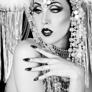 Puede incluir: Un retrato en blanco y negro de una mujer con un tocado y un collar de cuentas. Lleva un maquillaje de ojos oscuro y dramático y uñas largas y oscuras.