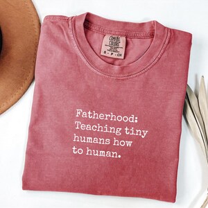 以下が含まれることがあります： 「Fatherhood: Teaching tiny humans how to human」の文字が入った赤みがかったオレンジ色のTシャツ。クルーネックで、首元に小さなタグが付いています。茶色の帽子と乾燥した葉が背景に一部写っています。