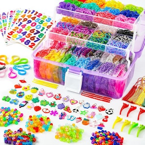 May include: A multi-tiered clear plastic storage case filled with assorted colorful rubber bands, accompanied by sheets of number and letter stickers, various small plastic charms, colorful beads, plastic hooks, and weaving tools.