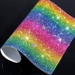 May include: A flexible sheet covered in a dense grid of small, reflective rhinestones arranged in a vibrant rainbow gradient. The surface transitions from blue and green to yellow, orange, and pink, creating a sparkling, multi-colored effect.