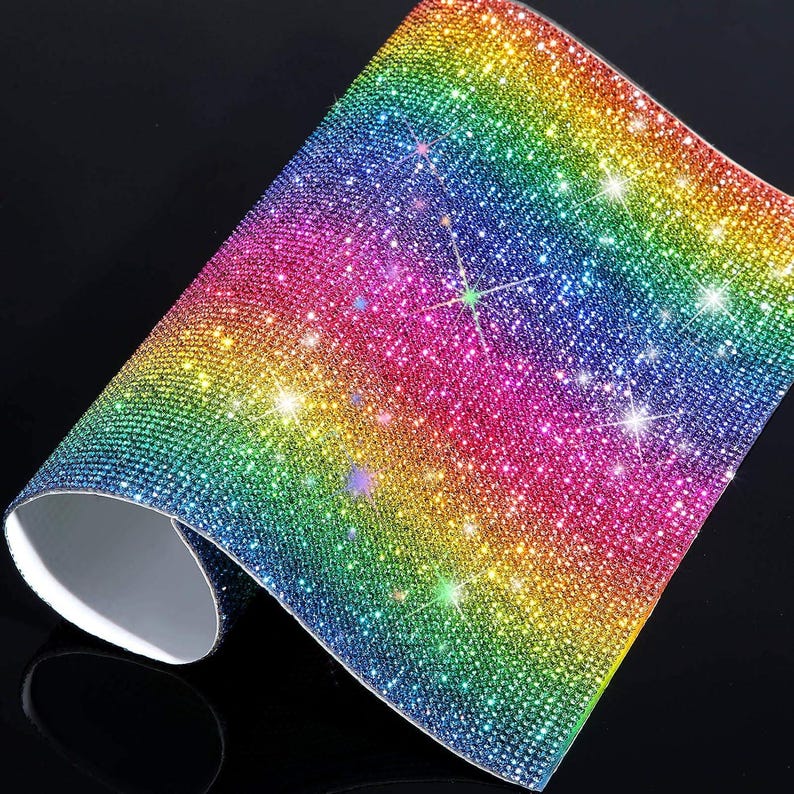 May include: A flexible sheet covered in a dense grid of small, reflective rhinestones arranged in a vibrant rainbow gradient. The surface transitions from blue and green to yellow, orange, and pink, creating a sparkling, multi-colored effect.