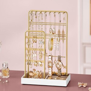 May include: A gold-colored jewelry organizer with multiple tiers and hooks for necklaces, earrings, and bracelets. The organizer also features a tray for rings and watches. The display is filled with various gold and pearl jewelry.