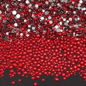May include: A close-up view showcases a dense collection of red rhinestones. The sparkling gems vary in size, with a mix of round and flat-backed shapes. The rhinestones are scattered across a dark surface, creating a vibrant and textured display.