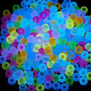 May include: A collection of colorful, glowing beads in various shapes and sizes. The beads are primarily cylindrical and donut-shaped, with a mix of vibrant colors including blue, green, yellow, pink, and purple. The beads are scattered against a black background.