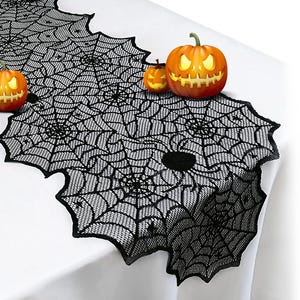 May include: A black lace table runner featuring a repeating spider web pattern with scalloped edges. The runner is decorated with two orange jack-o'-lanterns with glowing yellow eyes and mouths, positioned on top of the black fabric surface.