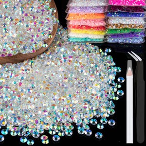 May include: A collection of iridescent rhinestones in various sizes and shapes, scattered on a black surface. Several small bags of colorful sequins are stacked in the background. A pair of tweezers and a white pencil are also present.