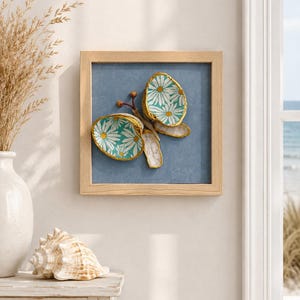 May include: A square framed wall art piece featuring a three-dimensional butterfly sculpture crafted from painted seashells. The butterfly wings are decorated with a teal and white daisy pattern and gold-painted edges, set against a blue background.