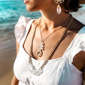 Pu&ograve; includere: Un orecchino a forma di fiore di loto color oro con un pendente in cristallo trasparente e una collana a strati composta da una catena dorata, una catenina sottile con pendente in cristallo e un ciondolo circolare, indossati con un top bianco a balze e un cappello di paglia.