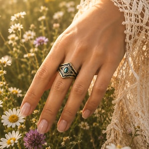 May include: A metallic ring featuring an oval-shaped, iridescent blue-green stone set within an ornate, darkened metal band with intricate etched detailing, worn on a finger amidst a field of white daisies and a white lace sleeve.