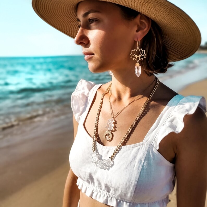 Pu&ograve; includere: Un orecchino a forma di fiore di loto color oro con un pendente in cristallo trasparente e una collana a strati composta da una catena dorata, una catenina sottile con pendente in cristallo e un ciondolo circolare, indossati con un top bianco a balze e un cappello di paglia.
