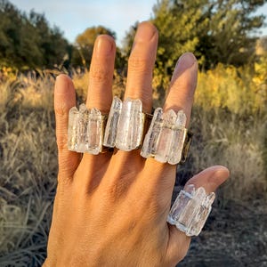 May include: Four clear crystal rings with gold-colored bands are displayed on a hand. The crystals have a rough, natural appearance. The background is blurred, with hints of green and brown, suggesting an outdoor setting. The rings are the focal point.