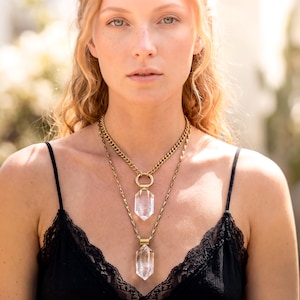 May include: A layered necklace set featuring two gold-toned chains of different lengths. Each chain holds a clear, hexagonal, faceted crystal pendant suspended from a gold-toned metal cap. The shorter chain is a curb link, while the longer is a twisted link.