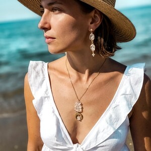 Pu&ograve; includere: Una collana e orecchini pendenti color oro con perline di cristallo trasparente sfaccettato e un piccolo ciondolo a forma di mezzaluna dorata. Il set di gioielli &egrave; indossato con un top bianco senza maniche con volant e un cappello di paglia.