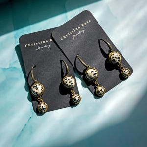 May include: Two pairs of dangle earrings with black and white dalmatian jasper stone accents. The earrings are displayed on black cards with the text "Christina Rose Jewelry".