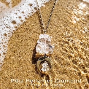 Pu&ograve; includere: Una collana con catena color bronzo, un grande cristallo grezzo trasparente e un ciondolo a mezzaluna che sostiene un cristallo grezzo pi&ugrave; piccolo. Il testo "Raw Herkimer Diamond Three Top Stones" &egrave; stampato nella parte inferiore.