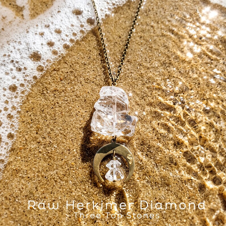 Pu&ograve; includere: Una collana con catena color bronzo, un grande cristallo grezzo trasparente e un ciondolo a mezzaluna che sostiene un cristallo grezzo pi&ugrave; piccolo. Il testo "Raw Herkimer Diamond Three Top Stones" &egrave; stampato nella parte inferiore.