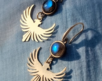 Puede incluir: Un par de pendientes colgantes con una piedra de labradorita azul iridiscente redonda engastada en metal oscuro, suspendida de un gancho. Debajo de cada piedra hay un dije de metal dorado con forma de ave en pleno vuelo.