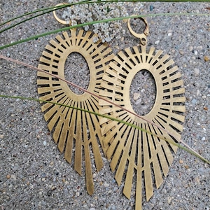 May include: A pair of large, gold-toned earrings with a sunburst design. The earrings are made of metal and have a geometric shape.