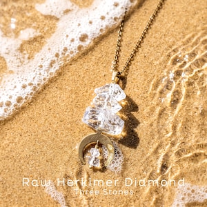 Pu&ograve; includere: Una collana color oro con tre cristalli di diamante Herkimer grezzi, trasparenti e sovrapposti, sospesi a una catenina sottile, con un ciondolo a forma di mezzaluna. Il testo "Raw Herkimer Diamond Three Stones" &egrave; visibile.