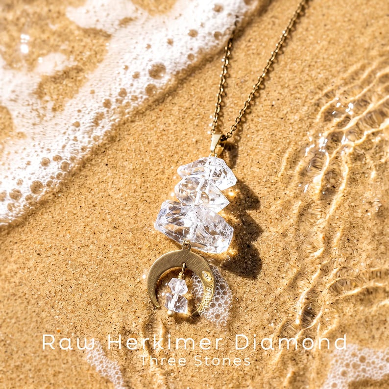 Pu&ograve; includere: Una collana color oro con tre cristalli di diamante Herkimer grezzi, trasparenti e sovrapposti, sospesi a una catenina sottile, con un ciondolo a forma di mezzaluna. Il testo "Raw Herkimer Diamond Three Stones" &egrave; visibile.