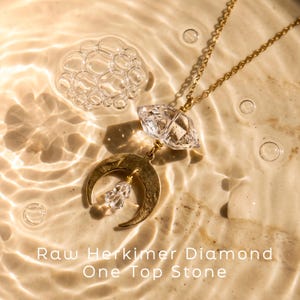 Pu&ograve; includere: Una collana color oro con un ciondolo di diamante Herkimer grezzo sospeso sopra un ciondolo a mezzaluna dorata, con un cristallo trasparente pi&ugrave; piccolo che pende dal centro. Il testo recita: Raw Herkimer Diamond One Top Stone.