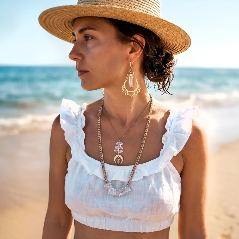 Pu&ograve; includere: Un cappello di paglia, orecchini dorati con montatura a forma di loto e punta di cristallo centrale, e una collana a strati con catena dorata, ciondolo a mezzaluna con cristallo e una fila di pietre di cristallo grezze su un top bianco.