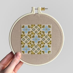 May include: A completed cross-stitch design in a round embroidery hoop. The pattern is geometric, using blue, olive green, and beige threads. The fabric is beige, and the hoop is off-white. The design is held in a 15 cm diameter hoop.
