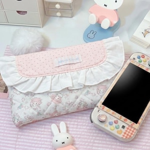 May include: A quilted fabric pouch featuring a pink polka dot flap with a white ruffle trim and a label reading APink Cloud. The main body has a white floral pattern with cartoon characters. Beside it is a handheld gaming console with a patterned skin.