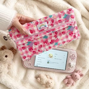 Puede incluir: Un estuche con estampado de cuadros vichy en rosa y blanco, decorado con fresas y una pequeña etiqueta rectangular con un arcoíris y la palabra HANDMADE. El estuche se sitúa sobre una consola de juegos portátil de color rosa claro.