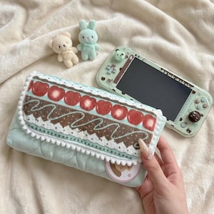 May include: A light mint green quilted carrying case with a flap featuring a pattern of red strawberries, brown chocolate icing, and white pom-pom trim. Beside it sits a matching mint green handheld gaming console with small animal thumb grips.