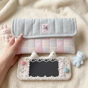 Op de afbeelding: Een gewatteerd tasje met een lichtgrijze klep en een pastelkleurig gestreept lichaam in roze, blauw en wit met een klein rechthoekig label. Daaronder een witte handheld gameconsole versierd met witte kanten randen, parels en kattenoortjes.