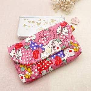 Puede incluir: Cartera de tela rosa con cierre de solapa, con un estampado de fresas, personajes de dibujos animados y lunares. Un botón blanco asegura la solapa. Una consola de juegos portátil blanca con siluetas doradas de Mickey Mouse está en el fondo.