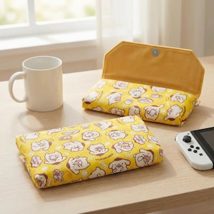 May include: Yellow fabric case with a cartoon dog pattern, open to reveal a yellow interior. A white mug and a game console are on the table. The case has a magnetic closure.