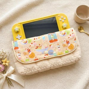 May include: A yellow handheld gaming console tucked into a cream-colored quilted fabric pouch. The pouch features a flap with a colorful print of various desserts and drinks. The console has two star-shaped joystick covers attached.