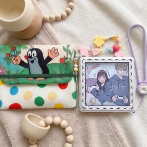 May include: A rectangular pouch featuring a cartoon mole character, a white base with colourful polka dots, and a green elastic band. Beside it is a square digital clock displaying an anime-style illustration of two people gaming, with a purple wrist strap.