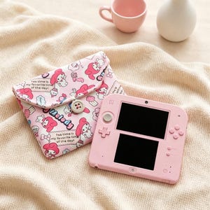 Puede incluir: Una consola de juegos portátil de color rosa claro con doble pantalla y una funda de tela a juego con un estampado de My Melody. La funda tiene un cierre de botón y muestra el texto 'Tea time is my favorite time of the day!' junto a varias ilustraciones del personaje.