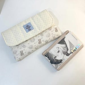 May include: A beige quilted case with a flap, featuring a small blue label and a pattern of teddy bears and other shapes. Beside it is a beige handheld gaming device with a black and white anime graphic.