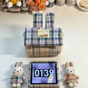Puede incluir: Un estuche de tela con estampado de cuadros azules y rosas, dos orejas de conejo erguidas y un parche rectangular con el texto miffy y una ilustración de un conejo con un tren. El estuche está rodeado por dos pequeñas muñecas de conejo.