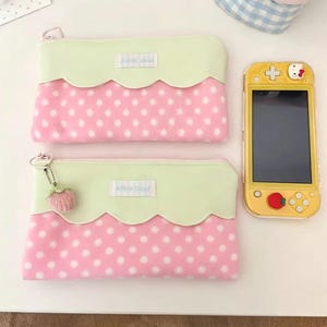 Puede incluir: Dos estuches rosas con lunares blancos, con la parte superior festoneada en verde claro y el texto "A Pink Cloud". Una consola de juegos portátil amarilla con una funda de Hello Kitty está al lado de los estuches. Un estuche tiene un dije de fresa rosa.