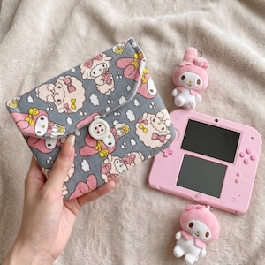 May include: A grey fabric pouch with a repeating pattern of pink and white cartoon characters, secured by a white button. Beside it sits a light pink handheld gaming console and two small plush character keychains with pink bows.