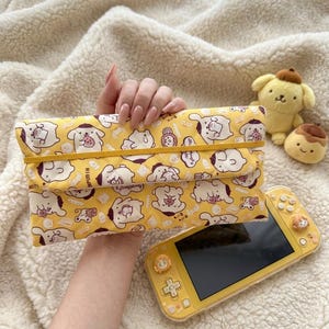 May include: A yellow quilted fabric pouch featuring a repeating pattern of the cartoon character Pompompurin. The rectangular pouch has a fold-over flap secured by a thin yellow elastic band. The design includes various illustrations of the character.