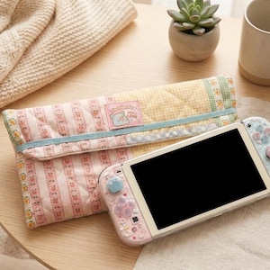 Puede incluir: Una consola Nintendo Switch con un diseño rosa pastel y azul, junto a una funda a juego. La funda está acolchada con un estampado floral y el texto "Sumikko Gurashi" es visible en una etiqueta.