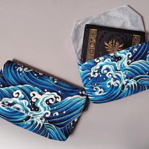 Op de afbeelding: Twee enveloppe-stijl tasjes met een levendig blauw en wit golfpatroon. Eén tasje is gesloten, terwijl de andere open is en een zwart elektronisch apparaat met gouden grafische details en een lichtgrijze voering aan de binnenzijde toont.