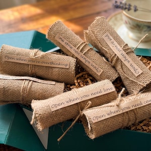 Könnte beinhalten: Eine tealfarbene Geschenkbox gefüllt mit gerollten Jutegarnrollen, die mit Bindfaden verschnürt sind. Jede Rolle hat ein Etikett mit Sätzen wie "Open when you're overthinking" und "Open when you need love". Eine Teetasse und ein Lavendelzweig sind im Hintergrund.