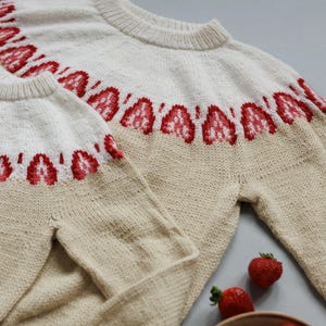 Peut inclure: Deux pulls en tricot beige avec un motif géométrique rouge et blanc autour du cou. Les pulls sont pliés sur une surface grise avec des fraises en arrière-plan.