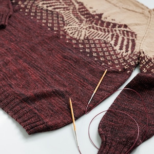 Puede incluir: Un suéter de punto marrón y beige con un patrón geométrico, colocado plano sobre una superficie blanca con dos agujas de tejer de madera y un ovillo de lana marrón.