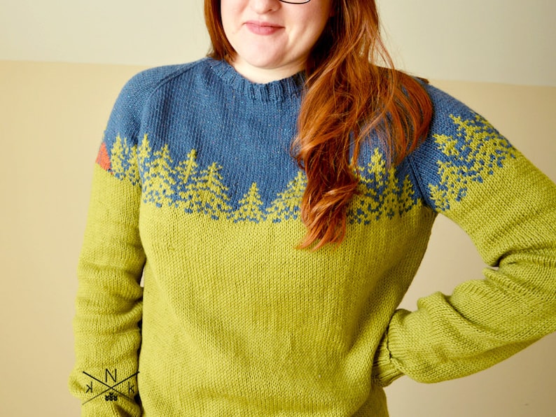May include: A green and blue knitted sweater with a forest scene design. The sweater has a crew neck and long sleeves.