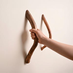 Może przedstawiać: Dwa drewniane wieszaki ścienne o naturalnym, zakrzywionym kształcie. Haczyki mają ciepły brązowy kolor i wyglądają na wykonane z gałęzi drzewa. Jeden haczyk jest trzymany przez dłoń. Haczyki są zamontowane na jasnobeżowej ścianie.