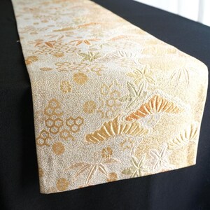 May include: A long, rectangular table runner featuring a cream and gold color palette. The fabric displays an intricate, raised woven pattern of stylized pine trees, bamboo leaves, and various floral motifs across the entire surface.