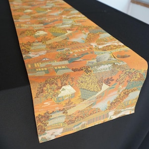 May include: A rectangular table runner featuring a vibrant orange background with a detailed, repeating pattern of traditional buildings, trees, and landscape elements in shades of green, blue, and gold, creating a complex, woven-style visual texture.